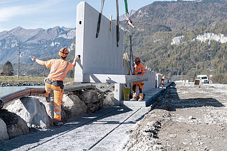 STRABAG - Mitarbeiter positionieren mithilfe eines Krans grosse Betonfertigteile auf einer Baustelle in alpiner Landschaft.