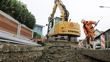 Liebherr-Bagger bei Aushubarbeiten in einer offenen Baugrube. Ein Bauarbeiter in orangefarbener Schutzkleidung säubert Grabenrand mit einem Besen. Im Hintergrund sind Werkleitungen gestapelt. Absperrungen und Verkehrsschilder sichern den Baustellenbereich.