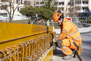 STRABAG-Bauführer in oranger Schutzkleidung prüft konzentriert die Eisenbewehrung an einer Holzschalung auf einer Baustelle.