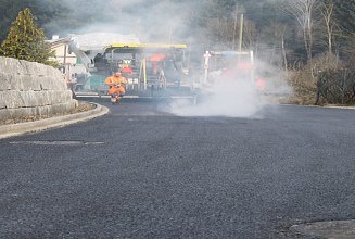 Ein Fertiger von Hinten mit dampfenden Asphalt auf dem die Bauarbeitenden die Maschine steuern und den Asphalt prüfen.