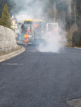 Ein Fertiger von Hinten mit dampfenden Asphalt auf dem die Bauarbeitenden die Maschine steuern und den Asphalt prüfen.
