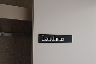 Nahaufnahme vom Türschild mit der Aufschrift «Landhaus» in weisser Schrift auf schwarzem Hintergrund, montiert an einer hellen Wand. Links im Bild ist ein Teil einer Garderobe mit Haken zu sehen.