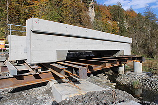 Ein grosses Betonfertigelement liegt auf provisorischen Stahlträgern über einem ausgehobenen Graben. Im Hintergrund stehen Bäume.