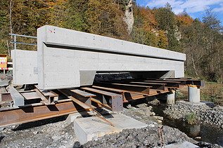 Ein grosses Betonfertigelement liegt auf provisorischen Stahlträgern über einem ausgehobenen Graben. Im Hintergrund stehen Bäume.