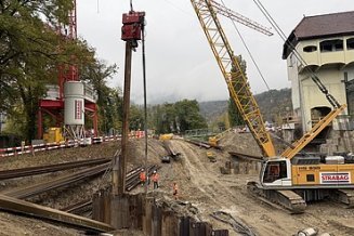 Luftbild: Baustelle am Fluss mit grossem rotem Kran, gelbem Bagger, Spundwänden und Baugrube neben einer Staumauer im Herbst.