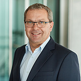 Technischer Gruppenleiter im Anzug lächelt in die Kamera. Im Hintergrund ist ein Lift zu sehen.
