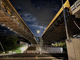  Ein Kran der STRABAG hebt nachts ein langes Brückenteil über Bahngleise. Die Baustelle ist hell erleuchtet.