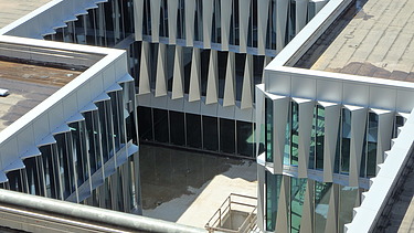 Blick von oben auf den Innenhof des modernen Neubaus mit markanter Fassade: Die Glasflächen sind durch dreieckig zulaufende, vertikal angeordnete Fassadenelemente rhythmisch strukturiert. Die Geometrie erzeugt ein prägnantes Licht- und Schattenspiel. Im Innenhof ist ein heller Betonboden mit einem abgesperrten Treppenabgang zu sehen.