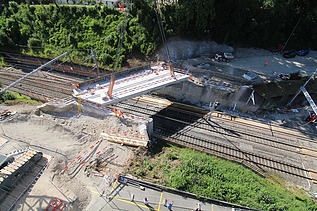 Eine Bahngleisüberquerung, die von einem Kran eingehoben wird. Im Hintergrund ist ein dichter Wald.