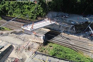 Eine Bahngleisüberquerung, die von einem Kran eingehoben wird. Im Hintergrund ist ein dichter Wald.