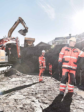 STRABAG - Mitarbeiter in oranger Schutzkleidung führen Vermessungsarbeiten auf einer Baustelle durch, im Hintergrund arbeitet ein Bagger bei Sonnenschein.