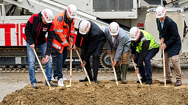 Mehrere Personen in Schutzkleidung und Helmen heben beim Spatenstich gemeinsam Erde aus. Im Hintergrund ist schweres Baustellengerät zu sehen.