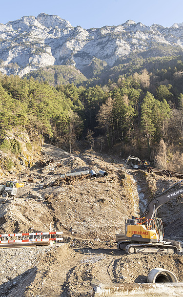 Ein Bagger der STRABAG räumt Geröll an einem steilen Hang unterhalb einer Felswand weg. Baustelle zur Hangsicherung im Gebirge.
