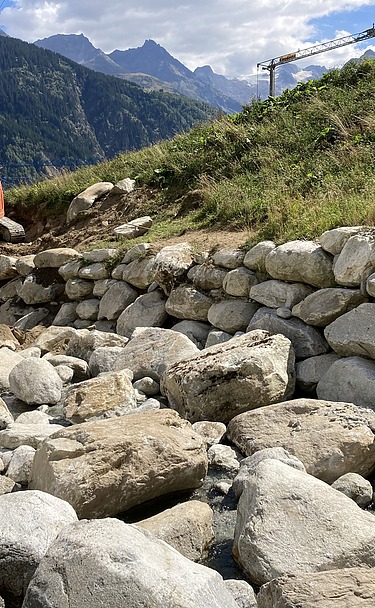 Schutz vor Naturgefahren - STRABAG Ingenieurbau STRABAG-Bagger baut Steinschutzwand in den Bergen am Aclettbach bei Disentis. Im Hintergrund ist eine Berglandschaft zu sehen.