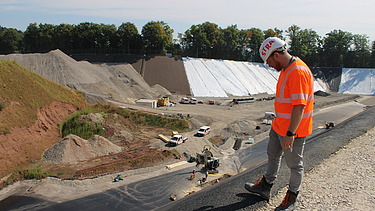 Bauarbeiter in orangefarbener Schutzkleidung steht am Rand einer steilen Böschung und blickt hinunter auf die Baustelle.