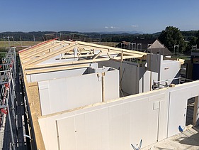 Rohbau der Labor-Erweiterung mit weissen vorgefertigten Wänden und sichtbaren Holzbalken für das Dach, eingerahmt von Baugerüsten, im Hintergrund ländliche Umgebung mit Feldern, Bäumen und Hügeln.