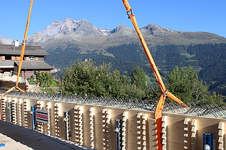 Ein langer Holzträger hängt an den Seilen eines Krans und wird über der Baustelle vor einer Berglandschaft positioniert.