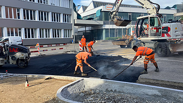  Ein Kran der STRABAG hebt an einem sonnigen Tag ein grosses Brückenelement. Im Hintergrund stehen mehrere Personen auf der Baustelle, neben einem Wohngebäude.