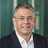 Einkauf's Gruppenleiter im Anzug lächelt in die Kamera. Im Hintergrund ist ein Lift zu sehen.