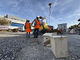 Zwei Schüler:innen warten vor dem Mini-Bagger in Baustellenweste und Baustellenhelme bis sie selbst ans Steuer dürfen