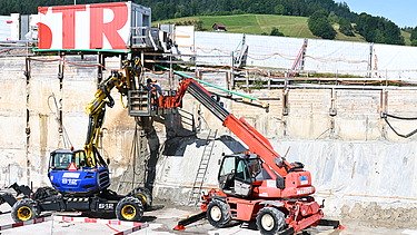 Zwei Baufahrzeuge in einer Baugrube bei der Arbeit. Im Hintergrund ein weisser STRABAG Container mit der Aufschrift STR in grossen roten Buchstaben.