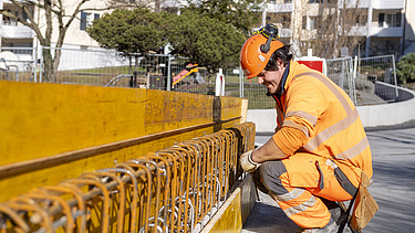 STRABAG-Bauführer in oranger Schutzkleidung prüft konzentriert die Eisenbewehrung an einer Holzschalung auf einer Baustelle.