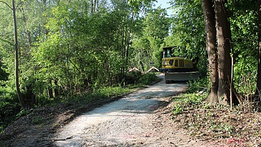 Ein gelber Bagger von STRAGAG steht auf einem geschotterten Waldweg, umgeben von dichten grünen Bäumen und Sträuchern.