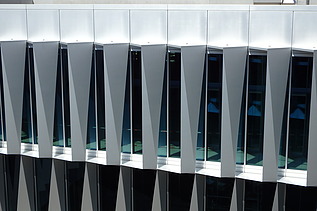 Blick von vorne auf die markante Fassade: Die Glasflächen sind durch dreieckig zulaufende, vertikal angeordnete Fassadenelemente rhythmisch strukturiert. Die Geometrie erzeugt ein prägnantes Licht- und Schattenspiel. 