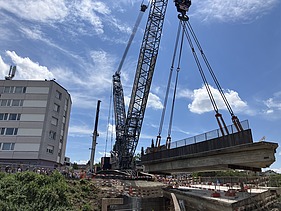  Ein Kran der STRABAG hebt an einem sonnigen Tag ein grosses Brückenelement. Im Hintergrund stehen mehrere Personen auf der Baustelle, neben einem Wohngebäude.