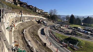  Panoramablick auf eine grosse Baustelle in Küssnacht. Eine Bohrpfahlwand stützt einen Hang mit Wohnhäusern und sichert die danebenliegende Strasse. Im Vordergrund sind Erdbewegungen und Baumaschinen zu sehen.