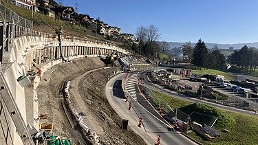  Panoramablick auf eine grosse Baustelle in Küssnacht. Eine Bohrpfahlwand stützt einen Hang mit Wohnhäusern und sichert die danebenliegende Strasse. Im Vordergrund sind Erdbewegungen und Baumaschinen zu sehen.