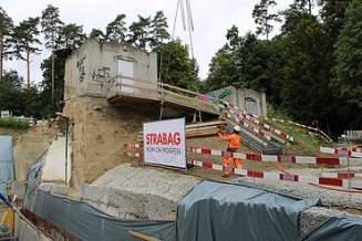 Bau eines Wasserreservoirs, im Vordergrund die Baugrube abgesperrt durch weiss/rote Latten. Im Hintergrund ein Bauarbeiter der gerade hilft Latten abzuladen, welche mit dem Kran über der Grube schweben. 