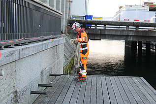 Sicherheitsbeauftragter knotet Seile an Ankerpunkte. Steht auf Holzsteg und im Hintergrund ist das Wasser der Limmat zu sehen.