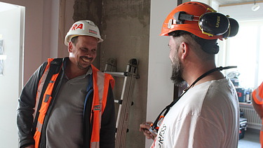 Zwei STRABAG Bauarbeiter in Helmen und Arbeitskleidung unterhalten sich lachend auf der Baustelle.
