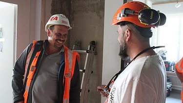 Bauführer im Gespräch - STRABAG Hochbau Zwei STRABAG Bauarbeiter in Helmen und Arbeitskleidung unterhalten sich lachend auf der Baustelle.