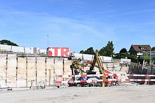 Mehrere Baufahrzeuge in einer Baugrube bei der Arbeit. Im Hintergrund ein weisser STRABAG Container mit der Aufschrift STR in grossen roten Buchstaben.