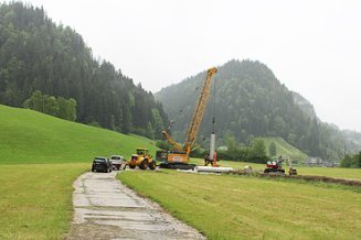 Baustelle im Grünen mit gelbem Kran, der einen Filterbrunnen für den Einbau aufstellt, und einem Bagger an einem Graben.