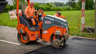 Ein Arbeiter fährt eine orange Strassenwalze und verdichtet frischen Asphalt auf einer Strasse.