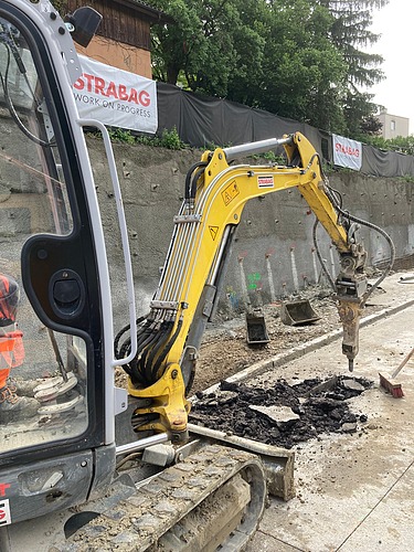 Eine kompakte Baumaschine spitzt Asphalt in einem Graben auf; daneben sind Baustellenmaterialien und Absperrungen sichtbar