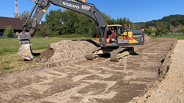  Ein Kran der STRABAG hebt an einem sonnigen Tag ein grosses Brückenelement. Im Hintergrund stehen mehrere Personen auf der Baustelle, neben einem Wohngebäude.
