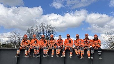  Ein Team von neun STRABAG Bauarbeiter:innen sitzt in orangen Uniformen für ein Gruppenfoto auf einer Brüstung unter blauem Himmel.