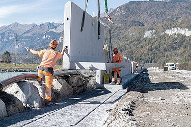 STRABAG - Mitarbeiter positionieren mithilfe eines Krans grosse Betonfertigteile auf einer Baustelle in alpiner Landschaft.