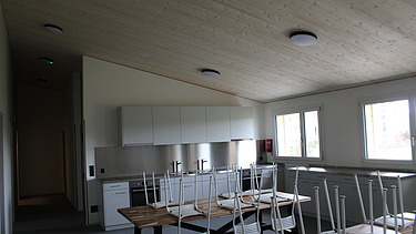 Heller Küchen- und Essraum mit weissen Schränken, grossen Tischen und gestapelten Stühlen.