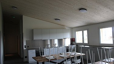 Heller Küchen- und Essraum mit weissen Schränken, grossen Tischen und gestapelten Stühlen.