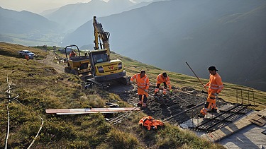 3 STRABAG-Bauarbeiter führen Handarbeiten an einem neuangelegten Weg aus. Im Hintergrund öffnet ein Bagger den Boden für den zukünftigen Weg und ein Tumber transportiert den Aushub ab. Im Hintergrund sieht man die Sonne durch eine Wolke durchstrahlen und eine Berglandschaft. 