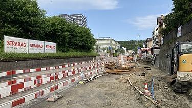 Eine Baustelle an der Strasse mit Sturzsicherung, Bauzäunen und verschiedenen Baugeräten mit einem leicht bewölkten Himmel.