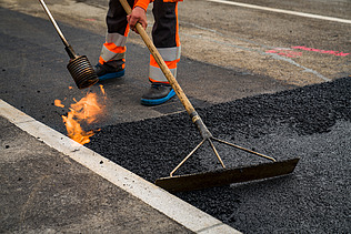 Ein Arbeiter glättet heissen Asphalt mit einem Werkzeug, daneben ist eine offene Flamme zu sehen.