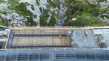 Blick von oben auf eine STRABAG Baustelle: Drei Bauarbeiter:innen giessen eine neue Strassenbrücke in den Schweizer Bergen.