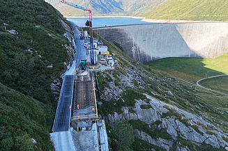 Baustelle von STRABAG in den Schweizer Bergen, mit einem Kran und einem in Bau befindlichen Strassenstück direkt neben einer Staumauer.