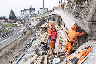 Zwei Bauarbeiter in leuchtenden Jacken inspizieren eine Bohrpfahlwand auf einer STRABAG Baustelle in Küssnacht. Im Hintergrund sind Absperrungen und ein Kran.